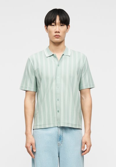 Homme portant une chemise à manches courtes vert clair à rayures verticales blanches et un jean bleu clair, debout devant un fond uni.