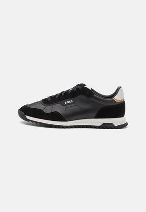 Zapatilla negra de caña baja con suela blanca, logo "BOSS" en el lateral, cordones negros y lengüeta trasera blanca con detalle en color tostado.