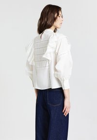 Blusa bianca con spalle arricciate, caratterizzata da delicati dettagli in pizzo, colletto alto e maniche voluminose. Abbinata a pantaloni in denim scuro.