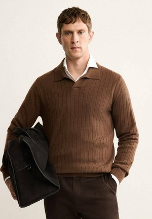 Uomo che indossa un maglione a coste marrone sopra una camicia bianca, con una borsa nera sotto il braccio, in piedi contro uno sfondo chiaro e semplice.