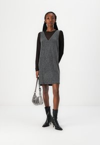 Vestuário cinza texturizado com decote em V, sobreposto a um top de malha preta. A modelo usa botas de salto pretas e segura uma bolsa prateada.