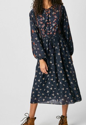 Kvinde iført en navyblå blomsterprintet mellemlang kjole med lange posede ærmer og brune snørebåndsankelstøvler.