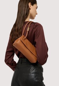 Bruine leren handtas met een gladde afwerking, met een gebogen vorm en een strap met gouden accenten. Gecombineerd met een bordeauxkleurig shirt en zwarte broek.