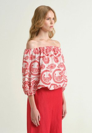 Femme blonde portant un chemisier blanc épaules dénudées avec des motifs rouges complexes et une longue jupe rouge, tournée vers la droite.