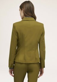 Blazer aderente verde oliva con una texture liscia e una silhouette strutturata; presenta un colletto a rever e cuciture posteriori sagomate.