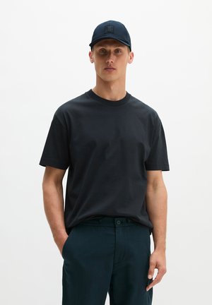 Man met een zwarte pet, zwart T-shirt met korte mouwen en donkere broek met één hand in de zak tegen een effen achtergrond.