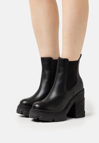 Nohy oblečené v černých kožených kotníkových botách s robustními platformovými podpatky a elastickými bočními panely na bílém pozadí.