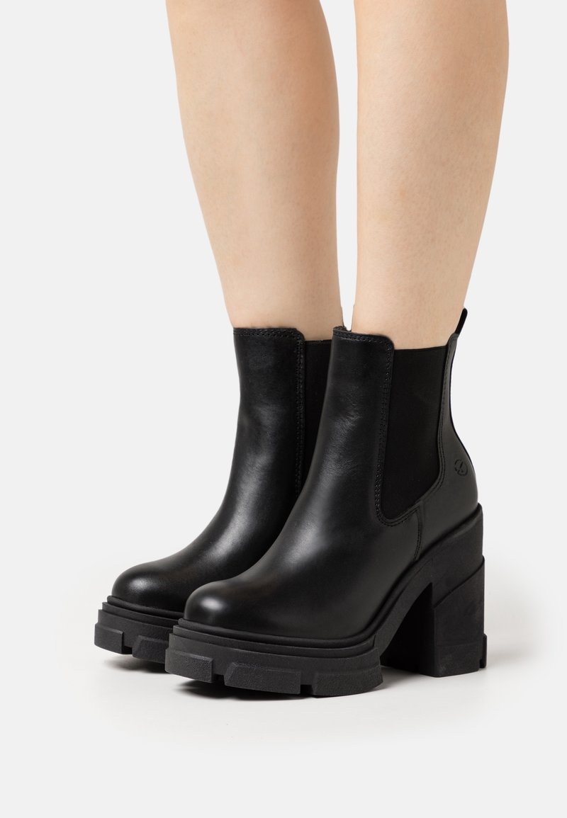 Nohy oblečené v černých kožených kotníkových botách s robustními platformovými podpatky a elastickými bočními panely na bílém pozadí.