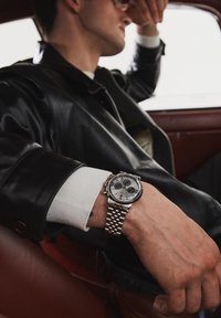 Montre chronographe en argent au poignet, avec un cadran blanc et des sous-cadrans bleus, bracelet en acier inoxydable, et une veste en cuir noir en arrière-plan.