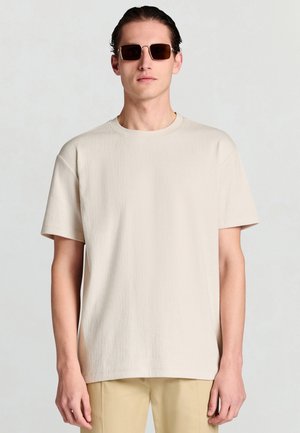 Mann mit dunkler Sonnenbrille, der ein schlichtes, beiges, strukturiertes Kurzarmhemd und hellbraune Hosen vor einem neutralen Hintergrund trägt.