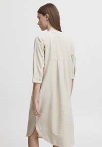 Robe beige, longue jusqu'aux genoux, en tissu léger, dotée de manches trois-quarts, d'un col rond et d'échancrures sur les côtés pour faciliter le mouvement.