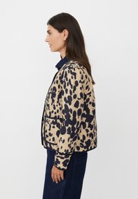 Giacca trapuntata beige con un motivo a macchie nere, caratterizzata da un colletto arrotondato, tasche frontali e dettagli in blu navy. Texture liscia.
