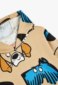 Sudadera color marrón claro con caras de perros de dibujos animados en negro, blanco, naranja y azul. El interior es de un color claro y tiene una textura suave.