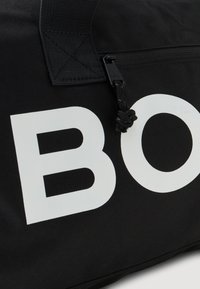 Črna potovalna torba iz trpežne tkanine; ima bel logotip, zadrgo in pletene ročaje z ojačanim šivanjem za podporo.