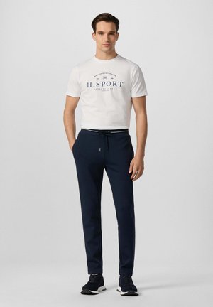 Jonge man staand, draagt een wit H. Sport T-shirt, marineblauwe broek met trekkoord en zwarte sneakers tegen een effen lichte achtergrond.