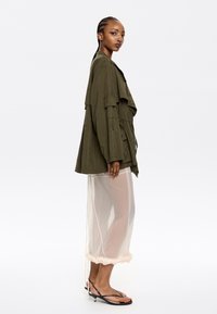Olive zelená oversize bunda s prednými vreckami a sťahovacím pásom, spárovaná s priehľadnou béžovou sukňou so volánmi a bočnými rozparky.