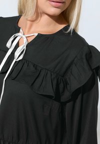 Lähikuva naisesta, jolla on päällään musta blusa, jossa on rypytyksiä rinnan ja hartioiden alueella, ja kauluksessa valkoinen kiinnitetty nauha.