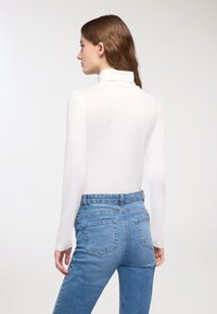 Biała bluzka z długim rękawem i stójką, zestawiona z dopasowanymi jasnoniebieskimi dżinsami. Charakteryzuje się gładką teksturą i klasycznym krojem. Widok z tyłu.