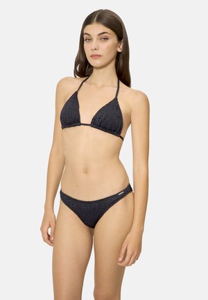 Mujer joven con cabello largo y oscuro, vestida con un bikini negro de dos piezas con textura, posando frente a un fondo liso y claro.