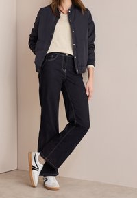 Veste de bombardier bleue marine, pull crème, jeans larges en denim foncé et baskets blanches avec accents noirs, présentés sur un fond neutre.