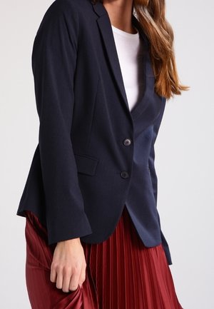 Blazer marine à deux boutons, avec des revers crantés et des poches latérales, associé à une jupe rouge plissée. Texture lisse, coupe ajustée.