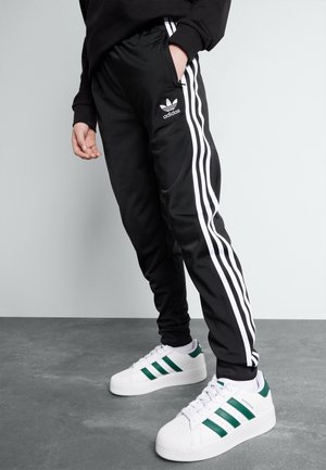 Person iført sorte Adidas træningsbukser med hvide striber og hvide Adidas sneakers med grønne striber, stående på en grå gulv.