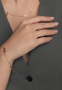 Złoty naszyjnik i bransoletka z motywem liści; bransoletka ma zielony akcent w postaci kulki, zaprezentowane na szarym tle materiału.