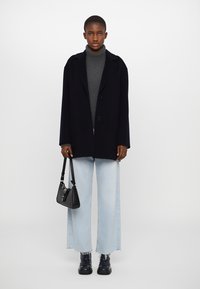 Manteau oversized bleu marine, col roulé gris, jeans larges bleu clair, sac à main noir et bottines brillantes noires. Textures lisses, sans motifs.