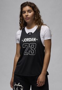 Svart baskettröja med "JORDAN" och "23" i vit text. Mesh-material, ärmlös design, lager-på-lager över en vit t-shirt.