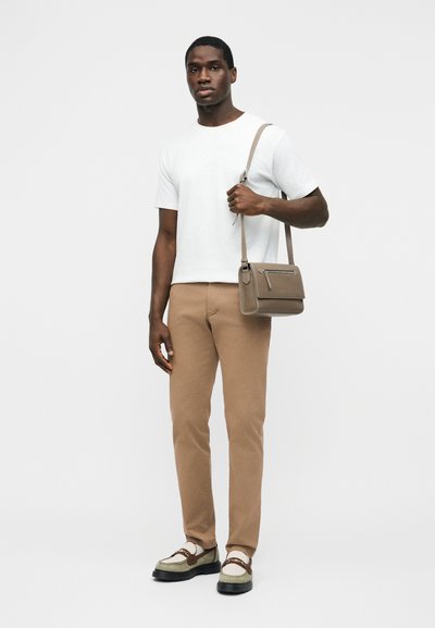 Homme portant une chemise blanche à manches courtes, pantalon beige, mocassins, tenant un sac bandoulière taupe sur un fond clair uni.