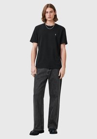 Camiseta de algodón negra con cuello redondo y mangas cortas, combinada con pantalones de mezclilla gris oscuro. Presenta un pequeño emblema en el pecho.