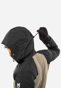 Veste imperméable noire et beige avec capuche ajustable, tissu texturé et poignets élastiques. Présente un logo sur le côté gauche.