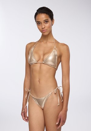 Bikini dorato metallico con reggiseno triangolare con lacci e hardware circolare, e lacci laterali regolabili sullo slip, su uno sfondo neutro.