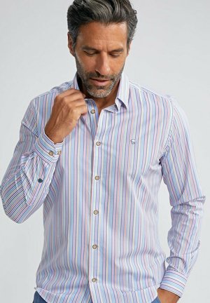 Homme ajustant le col d'une chemise à manches longues à rayures verticales multicolores et boutons marron sur un fond clair uni.