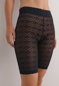Pantalones cortos de encaje negro con un intrincado patrón de diamantes y cintura con borde en forma de conchas. Hechos de material translúcido, que llegan hasta la mitad del muslo.