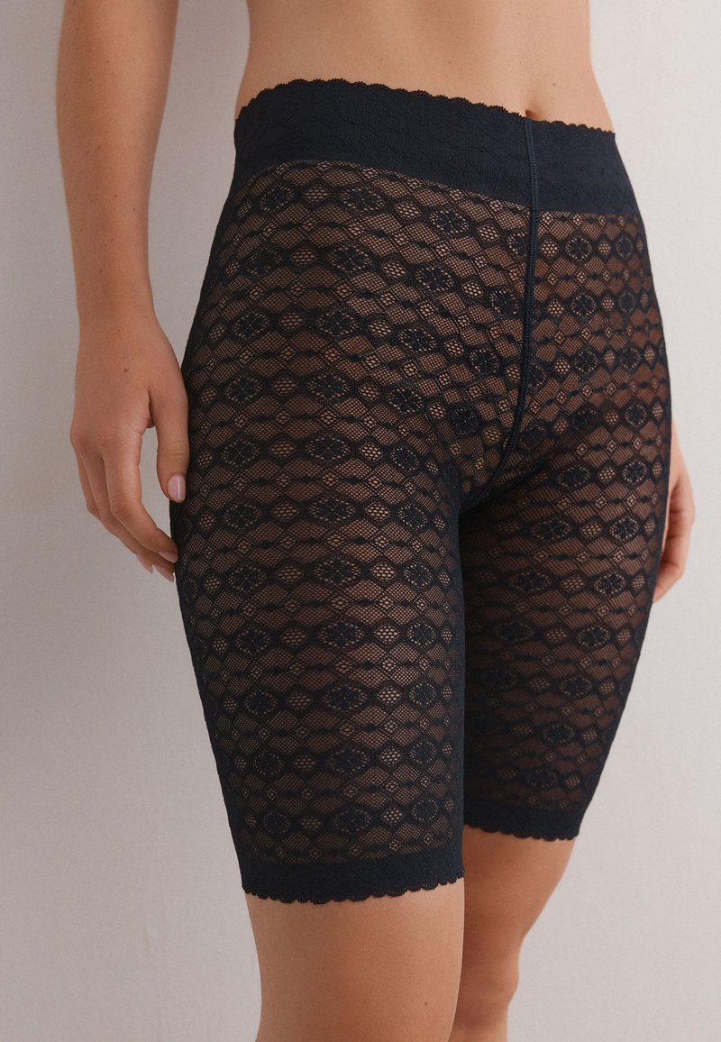 Pantalones cortos de encaje negro con un intrincado patrón de diamantes y cintura con borde en forma de conchas. Hechos de material translúcido, que llegan hasta la mitad del muslo.