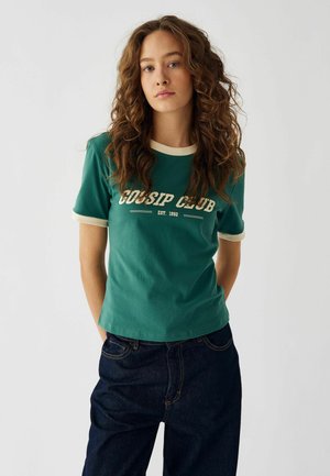 Grön bomullströja med korta ärmar, krämfärgad kant och tryckt text "GOSSIP CLUB EST. 1992." Kombinerad med mörka denimjeans.