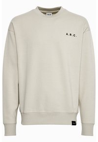 Beżowa bluza z długim rękawem, z okrągłym dekoltem, z czarnym napisem "A.R.C." na lewej piersi oraz małą czarną metką marki blisko dolnego brzegu.