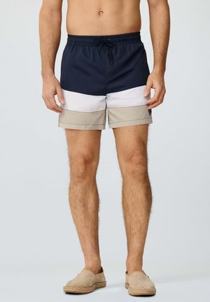 Torso y piernas de un hombre con bañador de bloques de color azul marino, blanco y beige, y zapatos deslizables tipo alpargatas beige sobre un fondo liso.