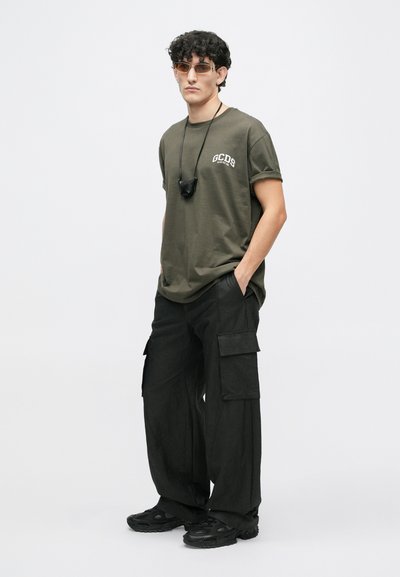 Olivno zelena bombažna majica s potiskom "GCDS", v kombinaciji s črnimi cargo hlačami s številnimi žepi in črnimi vezalnimi športnimi copati.