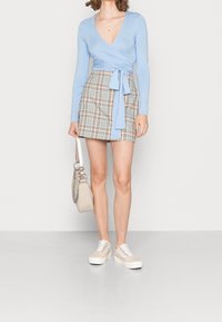 Hellblaues Wickeloberteil mit langen Ärmeln, kombiniert mit einem karierten Minirock in Beige, Rosa und Blautönen. Beigefarbene Sneakers und eine Umhängetasche.