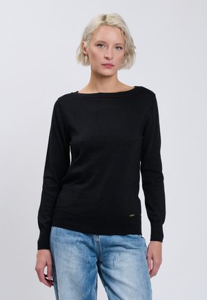 Blonde vrouw draagt een zwarte trui met lange mouwen en lichtblauwe spijkerbroek, staand tegen een effen lichtgrijze achtergrond.