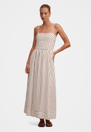 Nő, fehér midi ruhában, vékony barna függőleges csíkokkal, fodros mellrésszel, spagetti pántokkal, egy kézzel a csípőjén, szandálban áll.