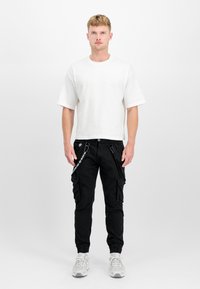Weißes Baumwoll-T-Shirt mit kurzen Ärmeln, kombiniert mit schwarzen Cargo-Hosen, die mehrere Taschen und verstellbare Seitengurte haben. Graue Sneakers.