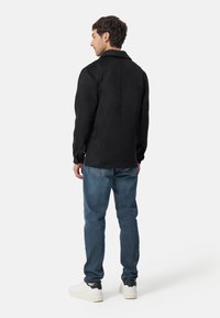 Zwarte suede jas met een rechte snit, voorzien van een kraag en lange mouwen, gedragen over blauwe jeans en witte sneakers. Gladtextuur.