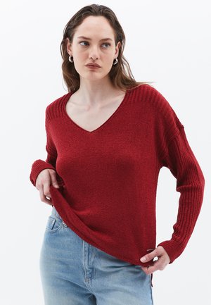Rød striktrøje med V-hals, ribstrikkede manchetter og kant. Stoffet har en tekstureret overflade. Parret med lyseblå jeans, der viser en afslappet pasform.