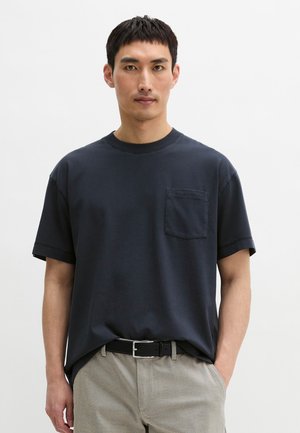 Mann trägt ein marineblaues kurzärmeliges T-Shirt mit Brusttasche und beiger Hose, steht vor einem schlichten weißen Hintergrund.
