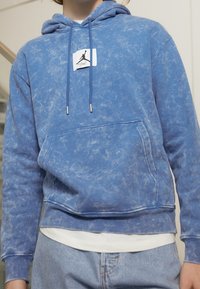 Sudadera azul de cuello redondo hecha de material suave, con bolsillo canguro, capucha con cordón y etiqueta de marca con un logotipo en silueta.