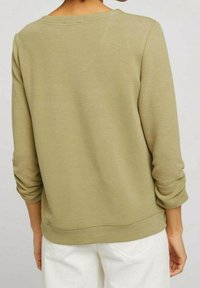 Maglione verde strutturato con scollatura rotonda e maniche a tre quarti, caratterizzato da una vestibilità rilassata e orlo a costine. Sotto si vedono dei pantaloni bianchi.