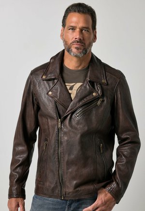 Mann mit kurzen dunklen Haaren und Bart, der eine dunkelbraune Lederjacke über einem grafischen T-Shirt vor einem schlicht grauen Hintergrund trägt.